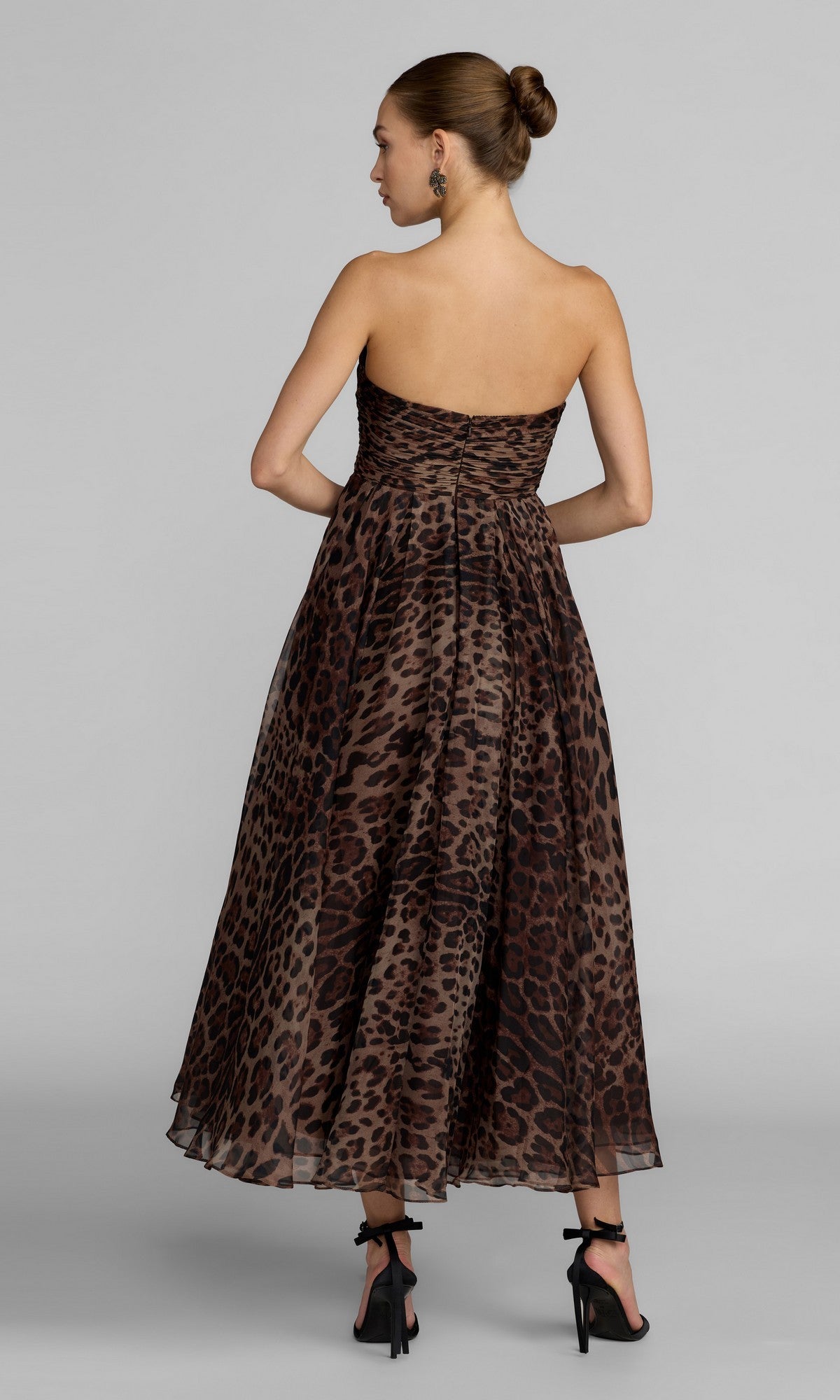 A woman wearing the Mac Duggal 56350 Strapless Leopard Formal Dress—an ankle-length, strapless gown with an a-line skirt—stands facing away in black high heels, her hair styled in a bun, against a plain light-colored background.
